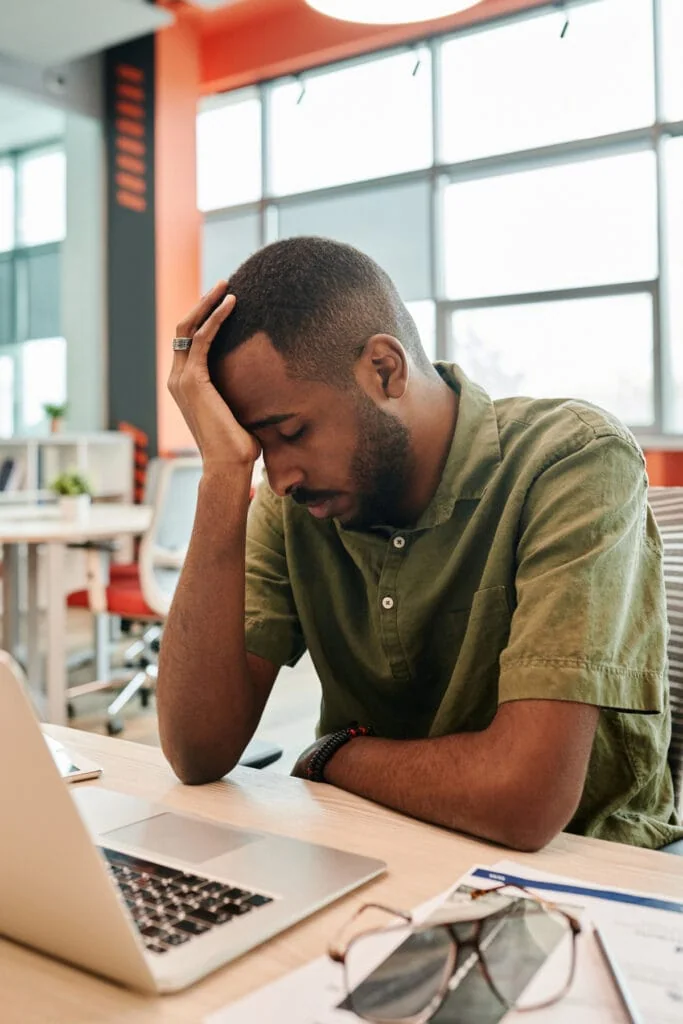 Man sitting at a desk with his head in his hand, looking exhausted in front of a laptop