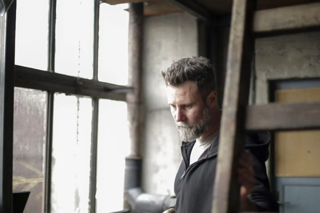 Man standing near a window in an industrial space, looking down in thought